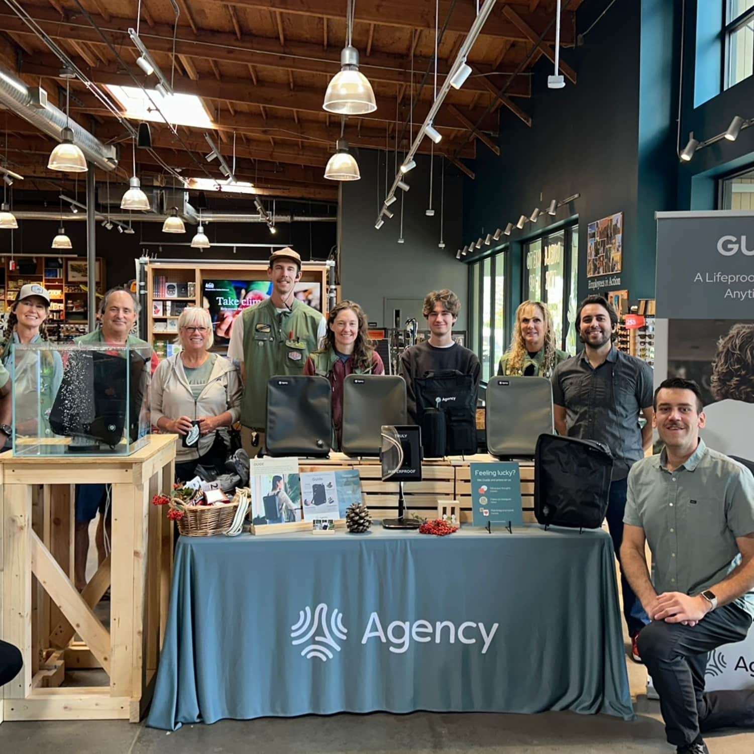 A mixed group of adults pose during a pop-up event at REI, behind a product showcase with branded bags, brochures, and informational signs.